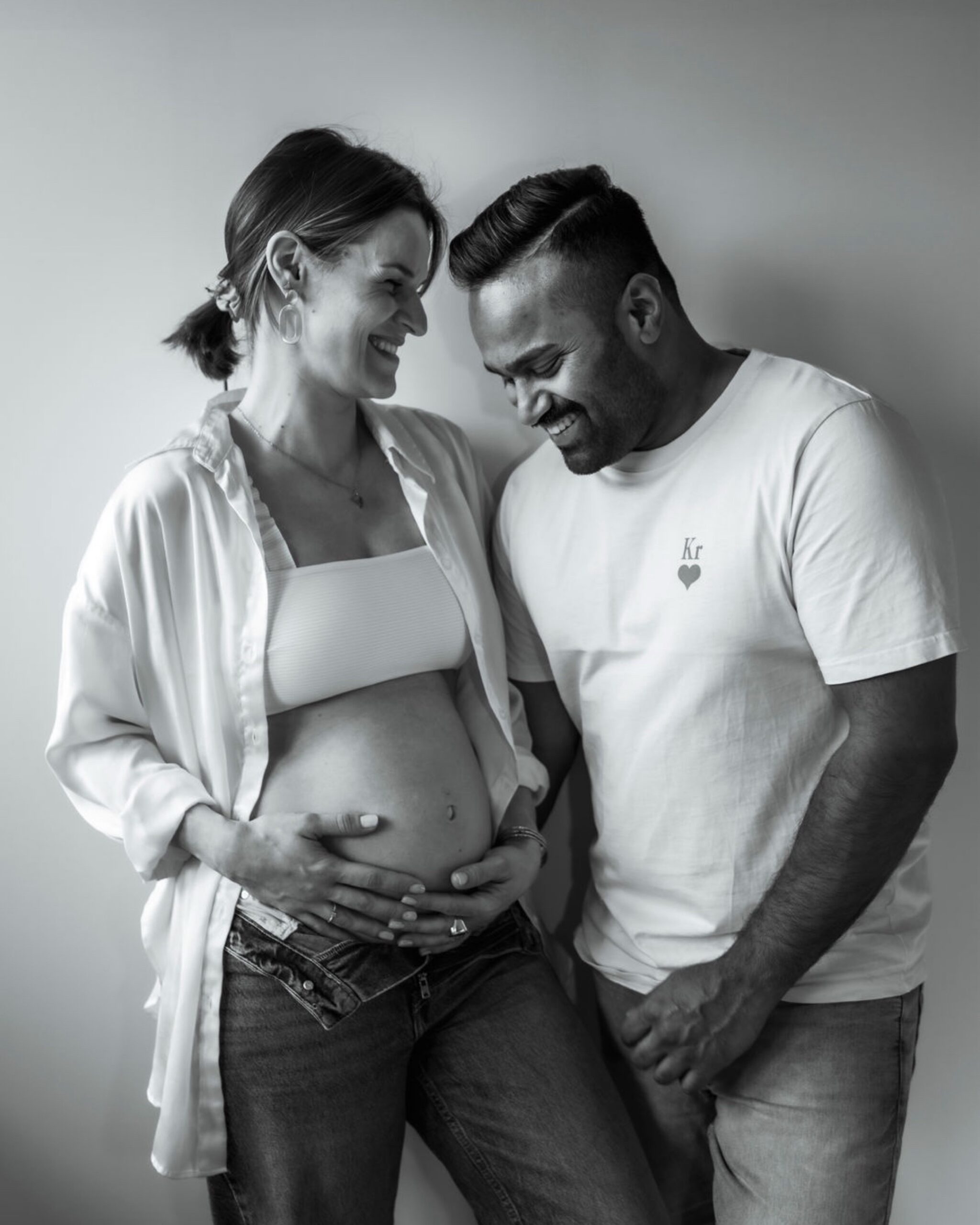 image0 Katharina lächelt Fabian an und hält ihren nackten Bauch mit ihren Händen. Fabian blickt lächelnd auf ihren Bauch und umarmt sie. Beide stehen vor einer Wand.