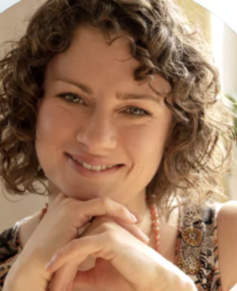 Bildschirmfoto 2025-06-24 um 14.44.17 Monika stützt ihr Kinn in ihre gefalteten Hände und blickt lächelnd in die Kamera. Es ist eine Nahaufnahme.