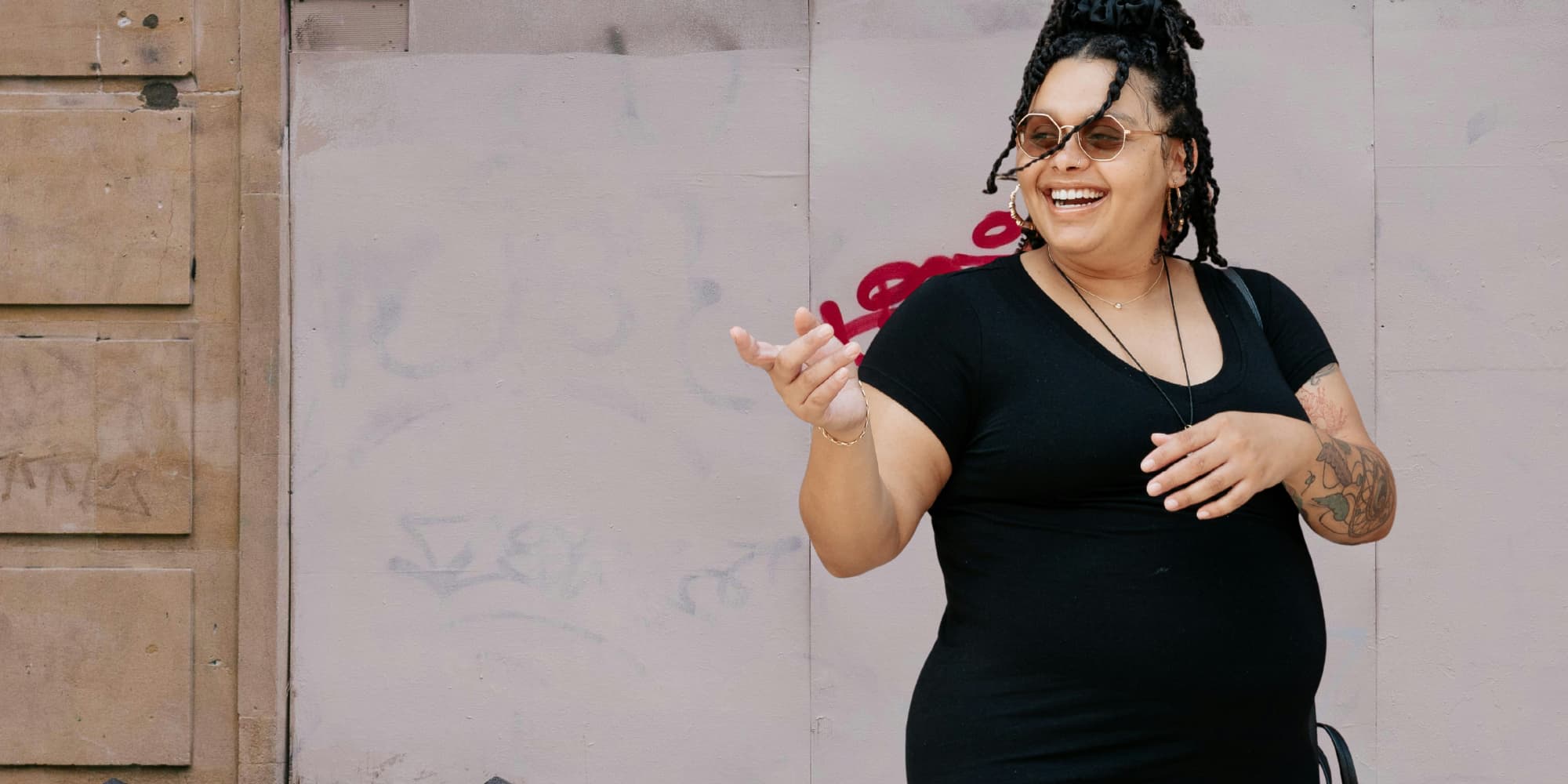 Schwangere Person überquert eine Strasse; sie lacht und hebt die Hand, um auf etwas zu zeigen, die andere Hand liegt auf ihrem Babybauch.